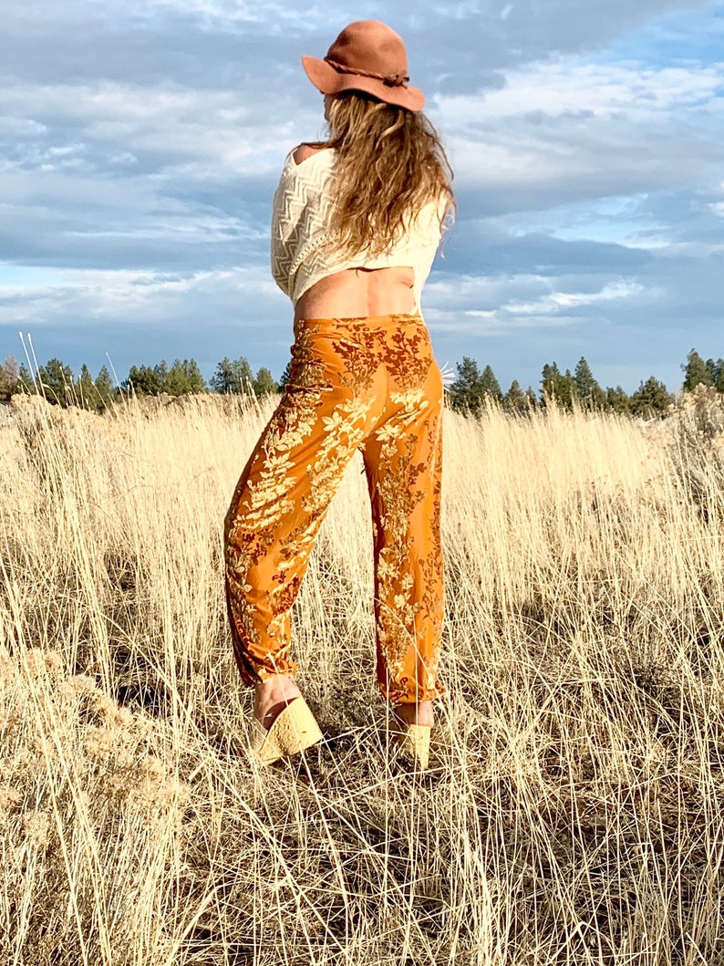 May include: A woman wearing a white sweater, brown hat, and orange floral print pants stands in a field of tall grass.