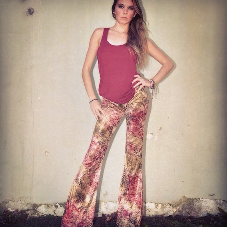 May include: A woman wearing a maroon tank top and brown and pink patterned bell bottom pants. She is standing in front of a light beige wall.