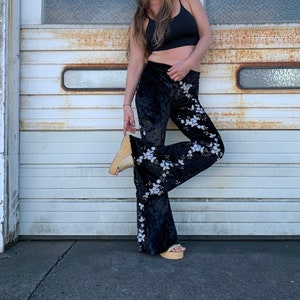 Pode incluir: Uma mulher vestindo um top preto curto, cal&ccedil;as de veludo preto boca de sino com padr&atilde;o floral, um chap&eacute;u de palha e &oacute;culos de sol. Ela est&aacute; em p&eacute; na frente de uma porta de garagem branca.