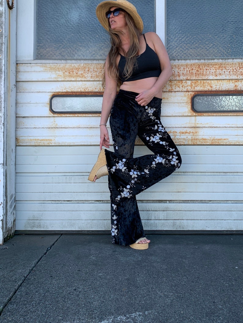 Pode incluir: Uma mulher vestindo um top preto curto, cal&ccedil;as de veludo preto boca de sino com padr&atilde;o floral, um chap&eacute;u de palha e &oacute;culos de sol. Ela est&aacute; em p&eacute; na frente de uma porta de garagem branca.