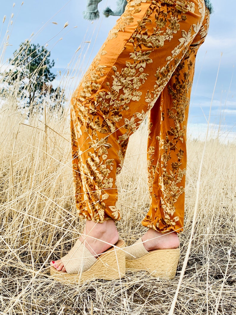 May include: A pair of brown platform sandals with a woven strap. The sandals are worn by a person standing in a field of tall, dry grass.