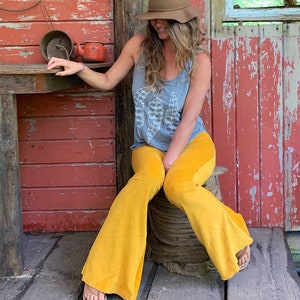 May include: A woman wearing a gray tank top with a white feather design, yellow bell bottom pants, and a brown fedora hat. She is sitting on a wooden spool in front of a red wooden wall.
