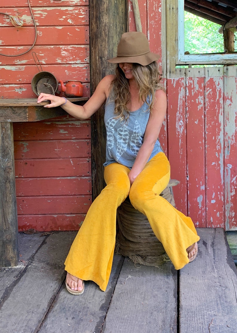 May include: A woman wearing a gray tank top with a white feather design, yellow bell bottom pants, and a brown fedora hat. She is sitting on a wooden spool in front of a red wooden wall.
