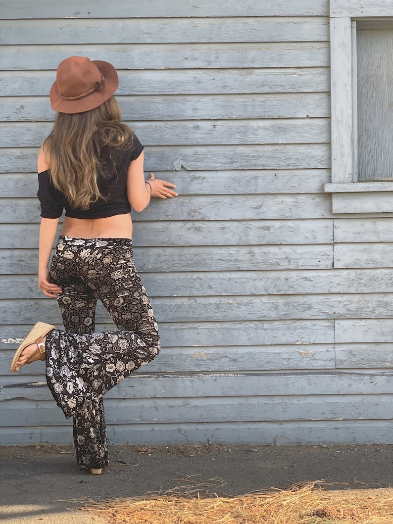 Op de afbeelding: Een vrouw draagt een bruine hoed, een zwart crop top en een zwarte en witte bloemenprint bellbottom broek. Ze staat voor een grijze houten muur.