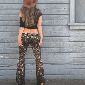 Op de afbeelding: Een vrouw draagt een zwart crop top en een zwarte en gouden bloemenprint bellbottom broek. Ze draagt een bruine fedora hoed en staat voor een grijze houten muur.