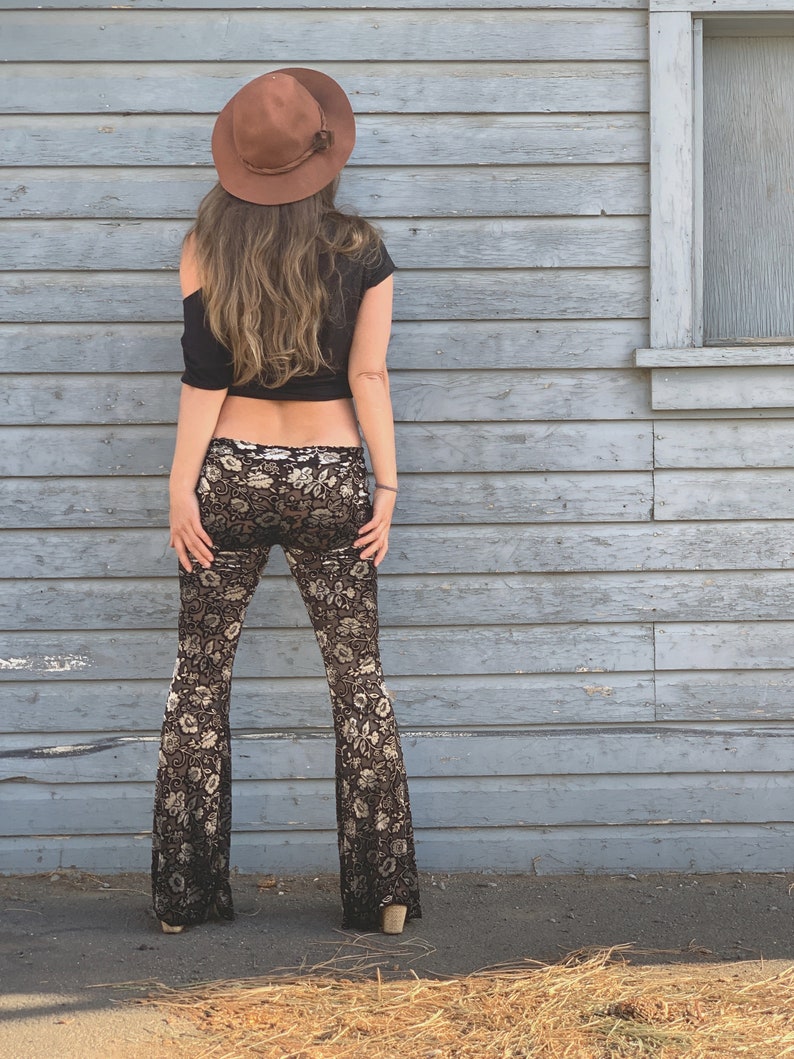 Op de afbeelding: Een vrouw draagt een zwart crop top en een zwarte en gouden bloemenprint bellbottom broek. Ze draagt een bruine fedora hoed en staat voor een grijze houten muur.