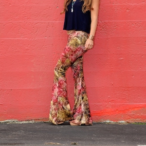 May include: A woman wearing a black tank top and patterned bell bottom pants stands in front of a red wall.