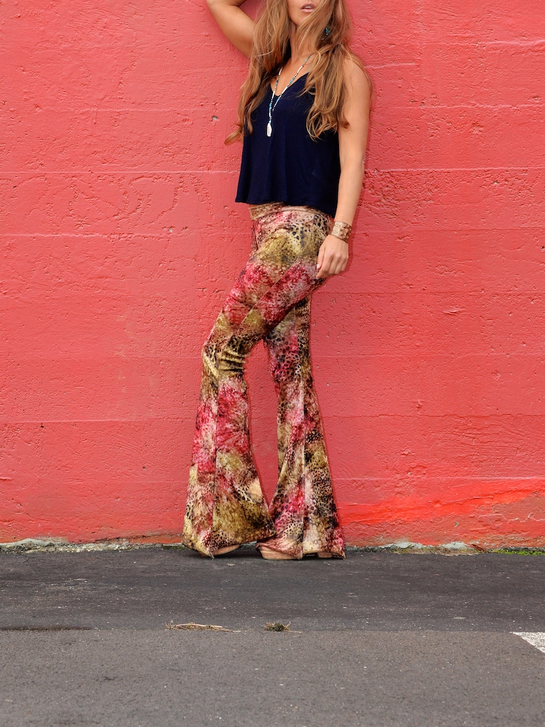 May include: A woman wearing a black tank top and patterned bell bottom pants stands in front of a red wall.