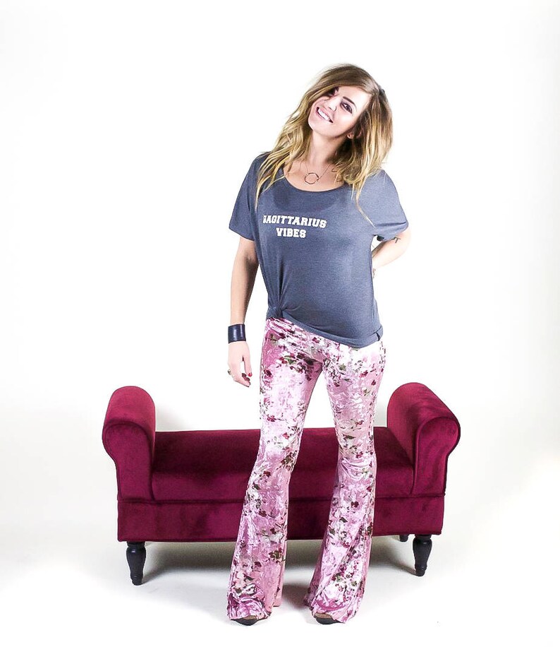 May include: A woman wearing a gray t-shirt with the text "Sagittarius Vibes" and pink floral bell bottom pants. She is standing in front of a small red velvet bench.