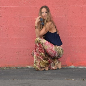 May include: A woman wearing a blue top and patterned pants sits with her legs crossed in front of a pink wall.