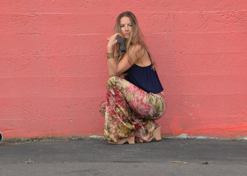 May include: A woman wearing a blue top and patterned pants sits with her legs crossed in front of a pink wall.