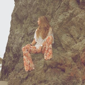 May include: A woman wearing a white crochet top and floral print pants sits on a large rock formation. The rock is a light brown color and has a rough texture.