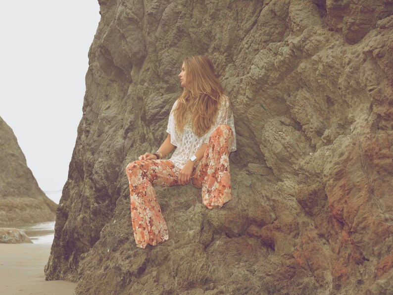 May include: A woman wearing a white crochet top and floral print pants sits on a large rock formation. The rock is a light brown color and has a rough texture.