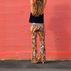 May include: A woman wearing a black top and brown and orange snake print pants stands in front of a red wall.