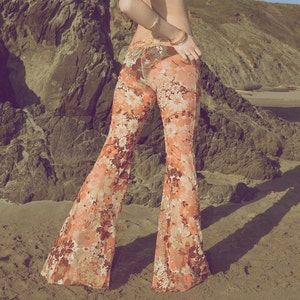 May include: A woman wearing a black bikini top and a pair of orange and white floral bell bottom pants. She is standing on a sandy beach with a rocky cliff in the background.