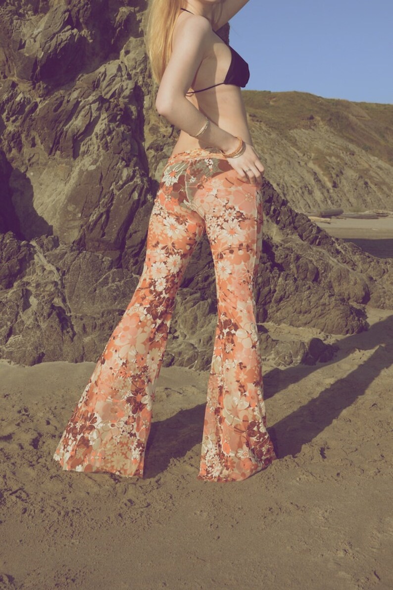 May include: A woman wearing a black bikini top and a pair of orange and white floral bell bottom pants. She is standing on a sandy beach with a rocky cliff in the background.