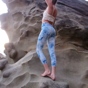 May include: A woman wearing a white crop top and blue and white tie-dye leggings stands on a large rock formation.