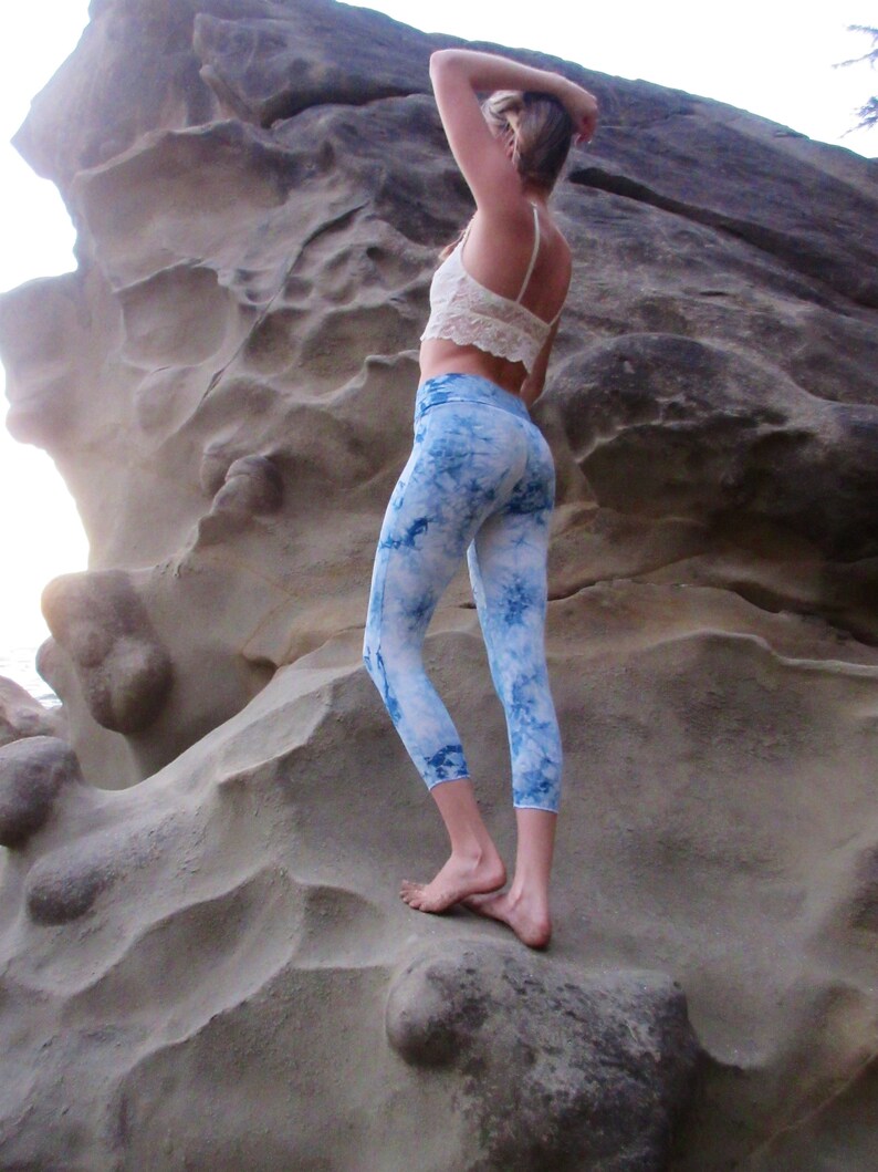 May include: A woman wearing a white crop top and blue and white tie-dye leggings stands on a large rock formation.
