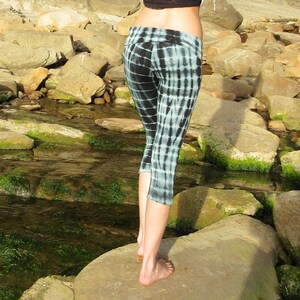 May include: A person wearing black and white tie-dye leggings with a black lace crop top. The person is standing on a large rock near a stream.