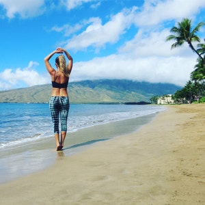 May include: A woman wearing a black sports bra and black and white plaid leggings stands on a sandy beach with a mountain range in the background. The ocean is in the foreground with waves lapping at the shore.