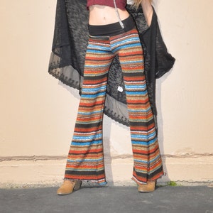 May include: A woman wearing a maroon crop top with a silver chain necklace, a black sheer cardigan, and striped bell bottom pants. She is standing on a black asphalt surface.