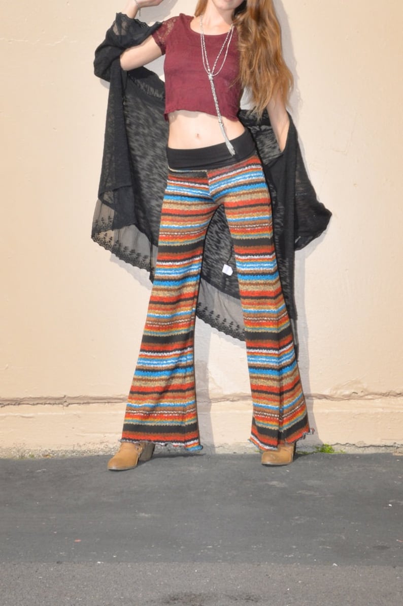 May include: A woman wearing a maroon crop top with a silver chain necklace, a black sheer cardigan, and striped bell bottom pants. She is standing on a black asphalt surface.