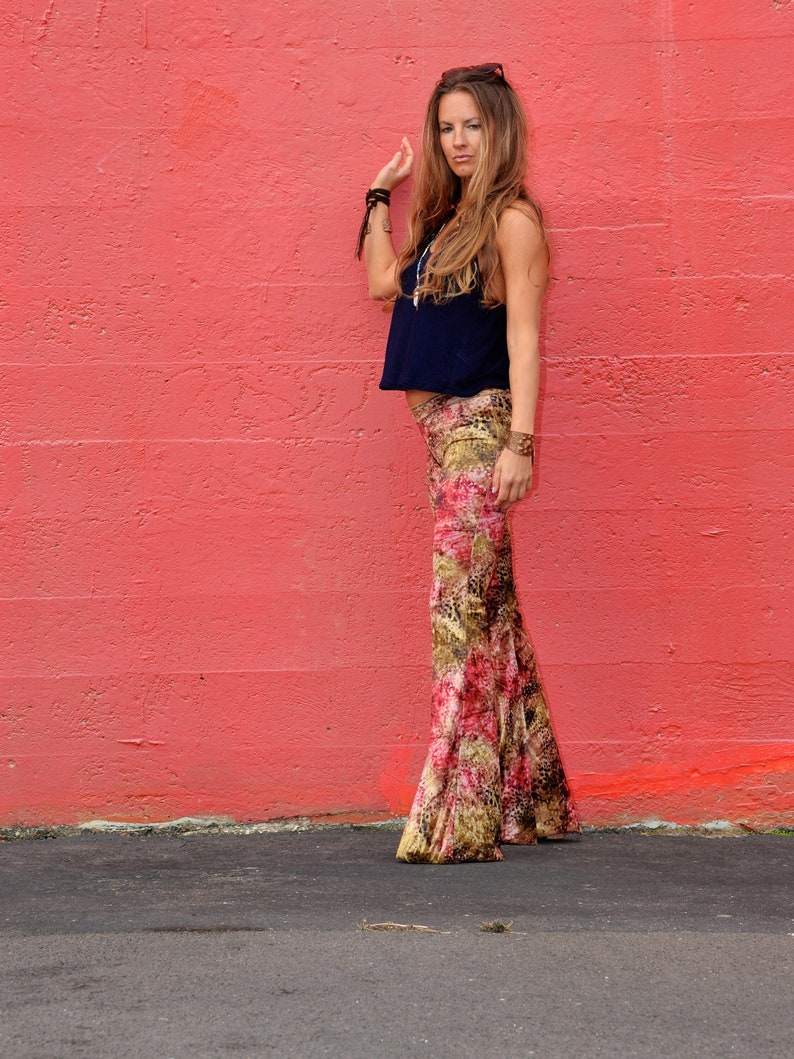 May include: A woman wearing a dark blue sleeveless top and patterned pants stands in front of a red wall. She is wearing a necklace and bracelets.