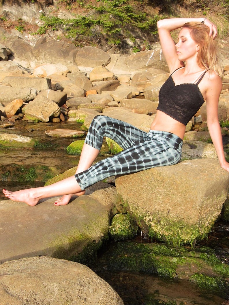 May include: A woman wearing a black crop top and black and white checkered pants sits on a large rock by a stream. She has her arm raised above her head and is looking off to the side.