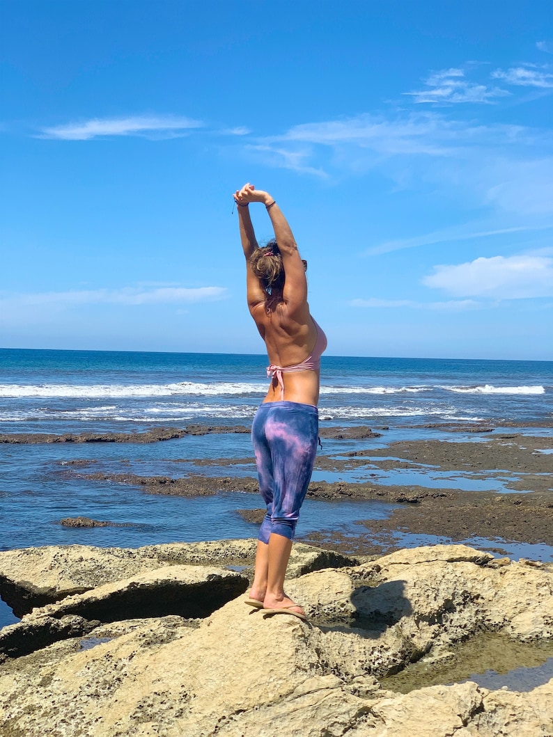 May include: A woman in a pink bikini top and blue and purple tie-dye leggings stretches her arms overhead while standing on a rocky beach.