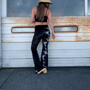Pode incluir: Cal&ccedil;a de veludo preto de boca de sino com bordado floral branco. A cal&ccedil;a est&aacute; sendo usada por uma pessoa que est&aacute; vestindo um top preto e um chap&eacute;u de palha.