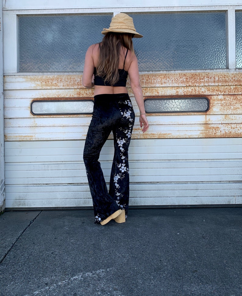 Pode incluir: Cal&ccedil;a de veludo preto de boca de sino com bordado floral branco. A cal&ccedil;a est&aacute; sendo usada por uma pessoa que est&aacute; vestindo um top preto e um chap&eacute;u de palha.