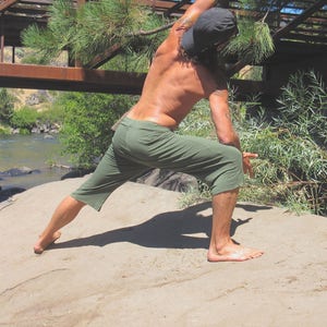 May include: A person wearing green pants and a black hat is stretching in a yoga pose on a sandy surface.