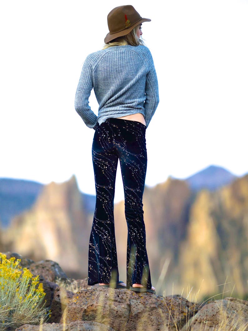 Puede incluir: Una mujer con un su&eacute;ter gris y pantalones acampanados azul oscuro con un estampado floral blanco. Lleva un sombrero marr&oacute;n con un adorno de plumas y est&aacute; de pie sobre una superficie rocosa.