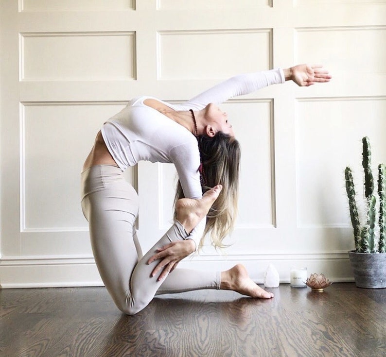 May include: A woman in a white long-sleeved shirt and beige leggings performs a yoga pose. She is kneeling with her right leg bent and her left leg extended behind her, her foot touching her right buttock. Her right arm is extended overhead, and her left arm is bent, her hand resting on her left thigh. Her hair is long and blonde. The background is a white wall with wainscoting.