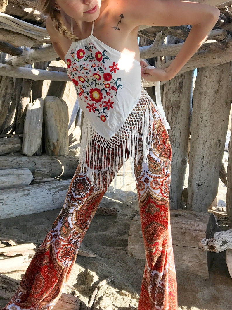 May include: A white halter top with red, yellow, and green floral embroidery and fringe trim. The top is paired with red, white, and gold paisley print bell bottom pants.