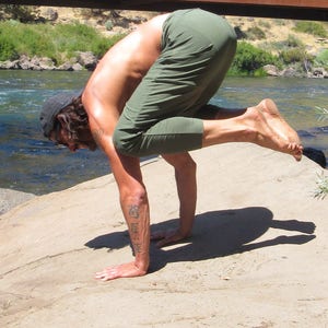 May include: A person performs a yoga pose outdoors, balancing on their hands with their legs raised in the air. They are wearing green trousers and have a tattoo on their arm.