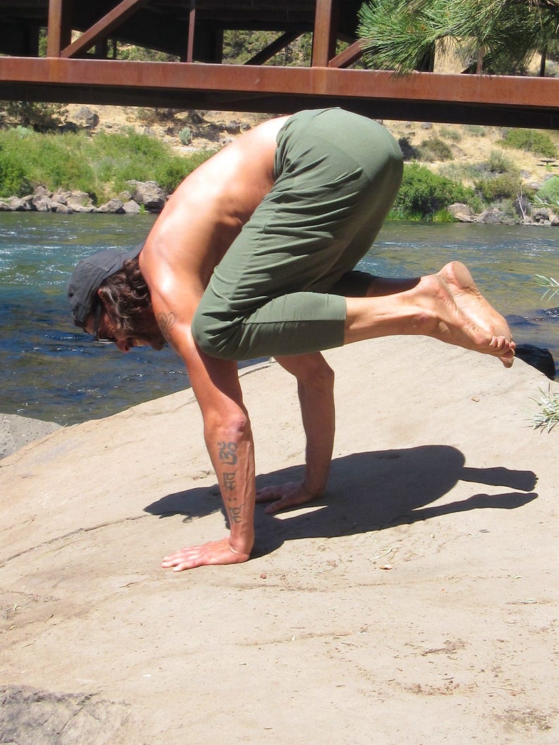 May include: A person performs a yoga pose outdoors, balancing on their hands with their legs raised in the air. They are wearing green trousers and have a tattoo on their arm.