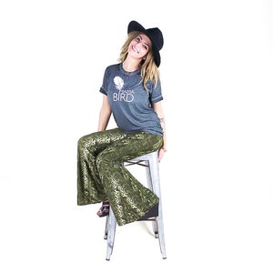 May include: A woman wearing a dark grey t-shirt with the text "Mama Bird" and a white feather graphic, black wide-brimmed hat, and green and gold snake print trousers sits on a silver stool.