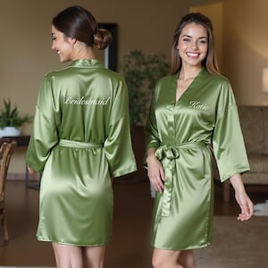 Two women wearing matching sage green satin personalized robes, one with “Bridesmaid” embroidered on the back and the other with “Katie” on the front. Elegant silk kimono dressing gowns perfect for bridesmaid robes, bridal party robes, and bride robes. Ideal for bridesmaid proposal gifts, maid of honor gifts, and mother of the bride or groom robes. Luxurious satin lounge and sleepwear, perfect for wedding morning getting-ready photos.