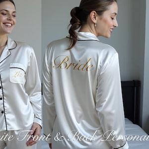 Two images showcase a woman wearing a luxurious ivory satin pajama set with elegant black piping. The front pocket is personalized with the name "Liz" in gold script, while the back features the word "Bride" in bold metallic gold. The smooth satin fabric adds a touch of elegance, making this set perfect for bridal party gifts, getting-ready moments, and personalized keepsakes. Ideal for weddings, bridesmaid proposals, and custom women’s sleepwear with front and back personalization options.