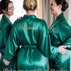 Two women in olive green satin personalized robes with gold embroidered names. Elegant bridal party robes for weddings, bridesmaid proposal gifts, and mother of the bride robes. Kimono-style silk dressing gowns perfect for lounge and sleepwear or maid of honor gifts.