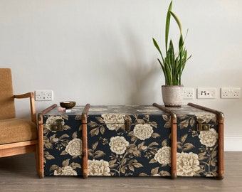 May include: A floral patterned vintage suitcase used as a coffee table with a brown armchair in the background.