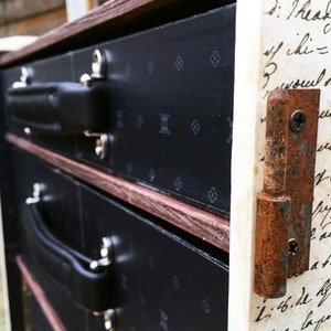 May include: A vintage wooden chest with three drawers, each with a black leather handle. The chest is decorated with a black and white pattern and has a rusty metal hinge on the right side. The side of the chest is covered with handwritten text.
