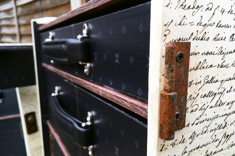 May include: A vintage wooden chest with three drawers, each with a black leather handle. The chest is decorated with a black and white pattern and has a rusty metal hinge on the right side. The side of the chest is covered with handwritten text.