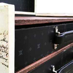 May include: A black wooden box with three drawers. Each drawer has a black leather handle and a black and white patterned front. The top of the box is made of wood and has a brown stain.