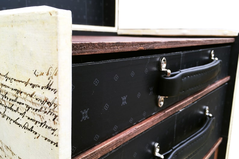 May include: A black wooden box with three drawers. Each drawer has a black leather handle and a black and white patterned front. The top of the box is made of wood and has a brown stain.