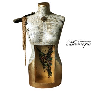 May include: A white and gold painted mannequin torso with a hummingbird illustration inside. The mannequin has a gold chain necklace draped over the shoulder and a lock and key detail on the chest. The text "by AM Florence Mannequins" is visible in the lower right corner.