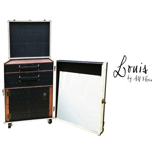 May include: A white vintage-style luggage cabinet with black and white patterned doors and drawers. The cabinet has a mirror on one side and a small shelf on the other. The cabinet is on wheels and has a brass handle. The text "Louis by AM Florence" is written on the cabinet.