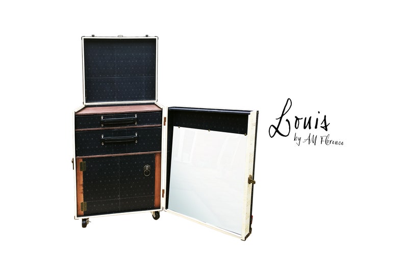May include: A white vintage-style luggage cabinet with black and white patterned doors and drawers. The cabinet has a mirror on one side and a small shelf on the other. The cabinet is on wheels and has a brass handle. The text "Louis by AM Florence" is written on the cabinet.