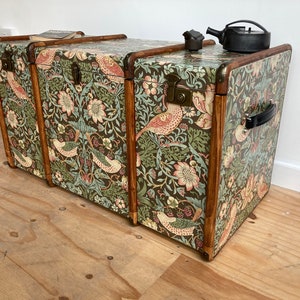 May include: Three vintage wooden storage trunks with a floral and bird patterned design. The trunks are stacked on top of each other and have brass hardware.